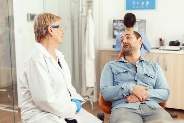 Stomatologue médecin principal discutant avec le patient avant d'examiner la santé bucco-dentaire alors qu'il était assis sur une chaise dentaire dans un bureau de stomatologie de l'hôpital