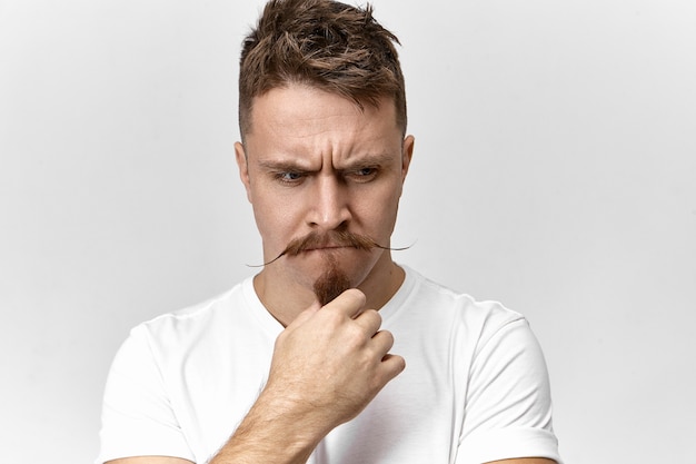 Stress, problèmes et émotions humaines négatives. Photo de beau jeune homme frustré en t-shirt blanc posant à l'intérieur, fronçant les sourcils et touchant sa barbe élégante, après avoir regardé inquiet inquiet