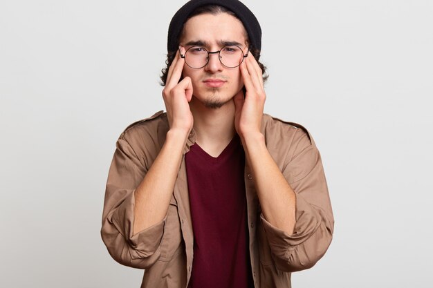 Stressé beau jeune toucher ses tempes avec les deux mains, ressentir des maux de tête, essayer de le réduire, semble bouleversé. Le modèle mince aux cheveux noirs porte un chapeau noir, des chemises rouges et beiges, des lunettes.