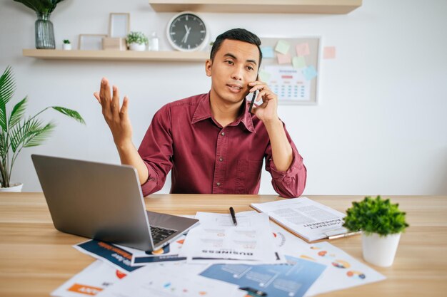 Stressé, un homme travaille au bureau avec son ordinateur portable, des papiers sur le bureau et un téléphone.