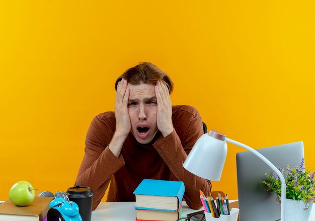 Stressé jeune étudiant garçon assis au bureau avec des outils scolaires mettant les mains sur les joues sur jaune