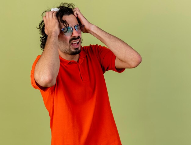 Stressé jeune homme malade portant des lunettes tenant une serviette en mettant les mains sur la tête avec les yeux fermés isolé sur un mur vert olive avec espace copie