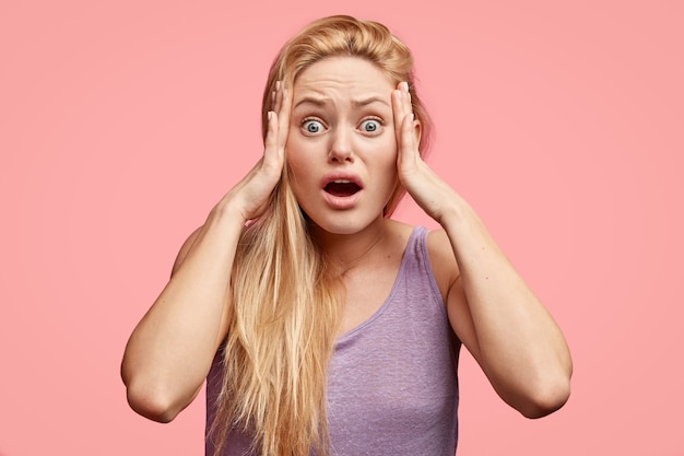 Stressée belle femme blonde garde les mains sur les tempes, a une expression effrayante, paniquée en entendant de terribles nouvelles, ouvre largement la bouche, vêtue d'un t-shirt violet, se tient seule contre un mur rose