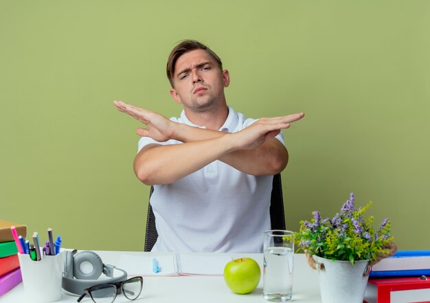 Strict jeune beau étudiant masculin assis au bureau avec des outils scolaires montrant le geste de non isolé sur vert olive