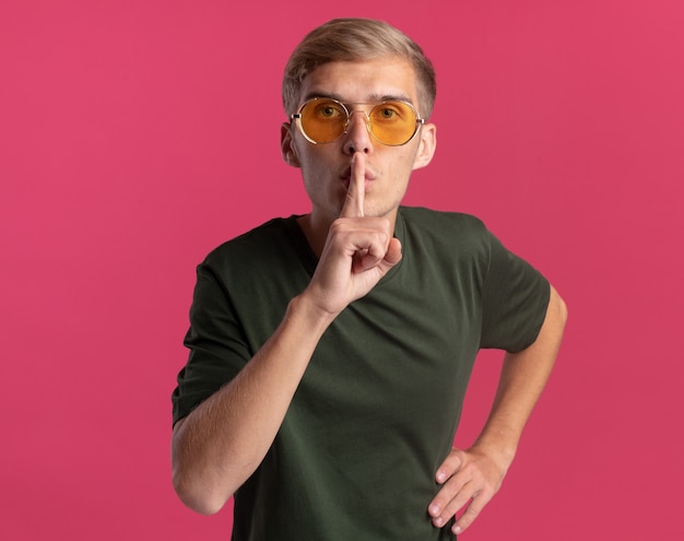 Strict jeune beau mec portant une chemise verte et des lunettes montrant un geste de silence mettant la main sur la hanche isolé sur un mur rose