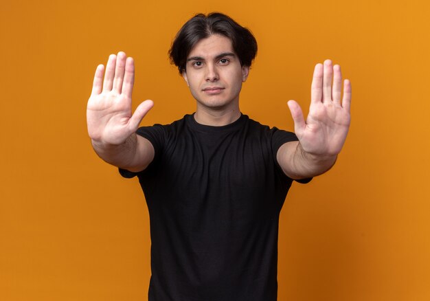 Strict jeune beau mec portant un t-shirt noir montrant le geste d'arrêt isolé sur un mur orange