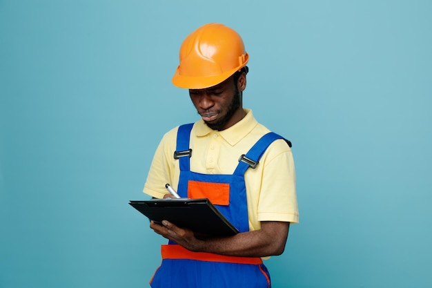 Strict jeune constructeur afro-américain en uniforme tenant et écrivant sur le presse-papiers isolé sur fond bleu