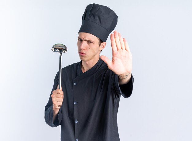 Strict jeune homme blond cuisinier en uniforme de chef et casquette tenant une louche regardant la caméra faisant un geste d'arrêt isolé sur un mur blanc avec espace de copie