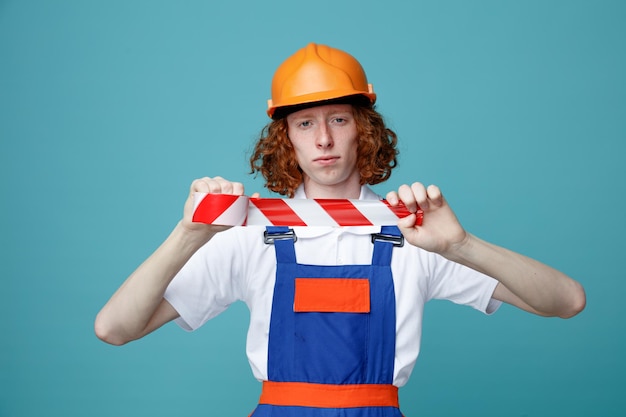 Strict Looking At Camera Jeune Constructeur Homme En Uniforme Tenant Du Ruban Adhésif Isolé Sur Fond Bleu