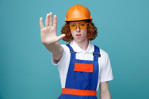 Strict montrant un geste d'arrêt jeune constructeur homme en uniforme isolé sur fond bleu