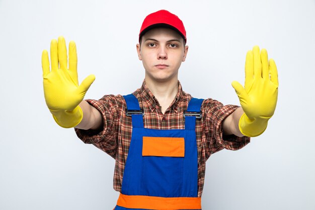 Strict montrant le geste d'arrêt jeune homme de nettoyage portant un uniforme et une casquette avec des gants