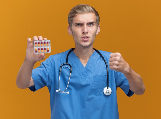 Strict young male doctor wearing doctor uniform with stethoscope holding pills isolé sur mur orange