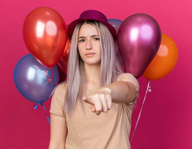 Stricte jeune belle fille portant un chapeau de fête debout devant des ballons vous montrant un geste