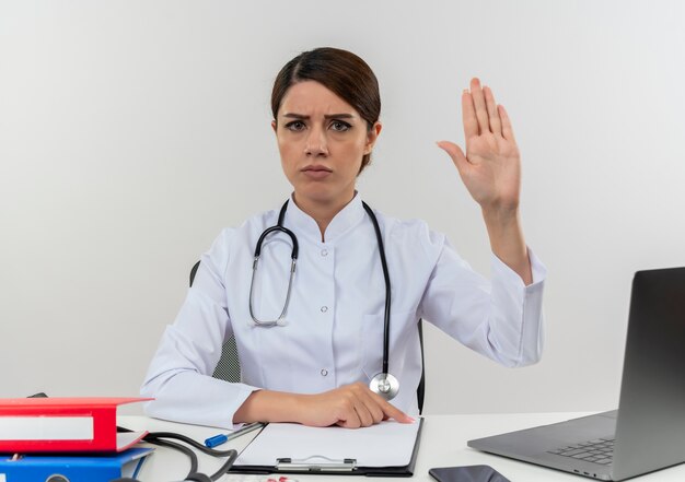 Stricte jeune femme médecin portant une robe médicale avec stéthoscope assis au bureau de travail sur ordinateur avec des outils médicaux montrant le geste d'arrêt avec copie espace