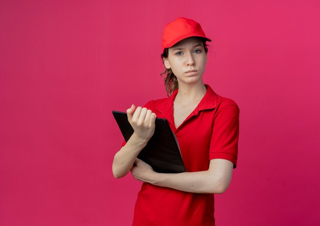 Stricte jeune jolie livreuse en uniforme rouge et cap holding presse-papiers regardant la caméra isolée sur fond cramoisi avec espace de copie