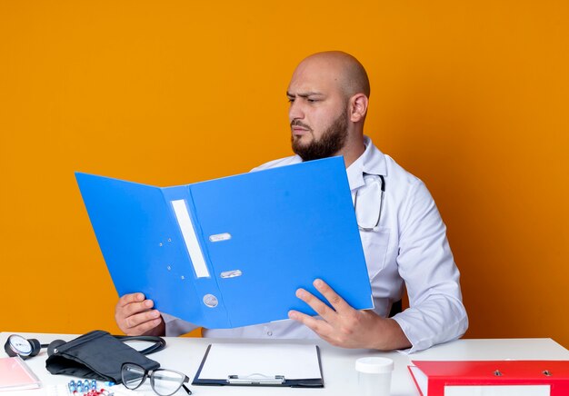 Stricte jeune médecin de sexe masculin chauve portant une robe médicale et un stéthoscope assis au bureau de travail avec des outils médicaux tenant et regardant le dossier isolé sur fond orange