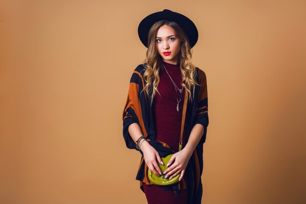 Studio gros plan portrait de jeune femme blonde fraîche en poncho de paille marron, chapeau à la mode noir en laine et lunettes rondes regardant la caméra. Le cuir vert avait un sac.