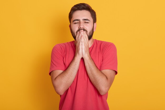 Studio jeune beau mec aux yeux fermés espère la fortune et la bonne chance, se tient vêtu d'un t-shirt décontracté rouge, garde les mains en prière, isolé sur jaune.