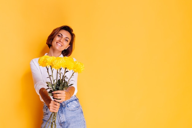 Studio tourné sur fond jaune heureux femme caucasienne cheveux courts portant des vêtements décontractés chemise blanche et pantalon en jean tenant bouquet d'asters jaunes