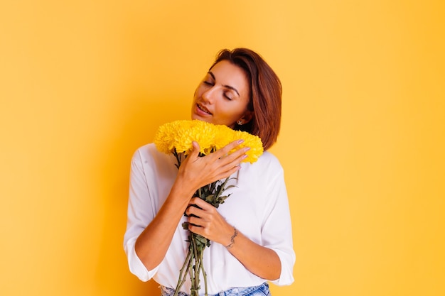 Studio tourné sur fond jaune heureux femme caucasienne cheveux courts portant des vêtements décontractés chemise blanche et pantalon en jean tenant bouquet d'asters jaunes