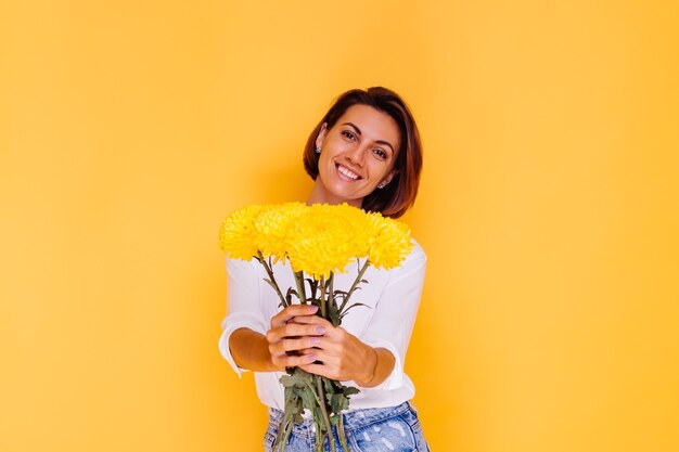 Studio tourné sur fond jaune heureux femme caucasienne cheveux courts portant des vêtements décontractés chemise blanche et pantalon en jean tenant bouquet d'asters jaunes