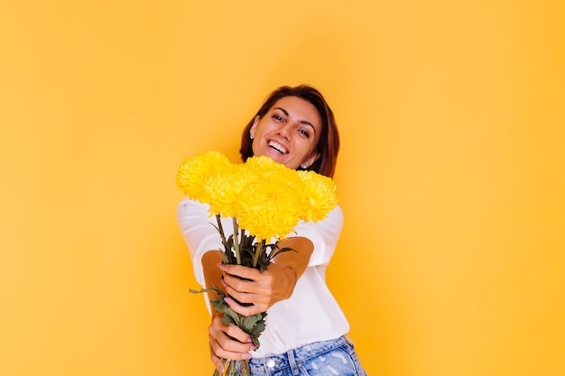 Studio tourné sur fond jaune heureux femme caucasienne cheveux courts portant des vêtements décontractés chemise blanche et pantalon en jean tenant bouquet d'asters jaunes