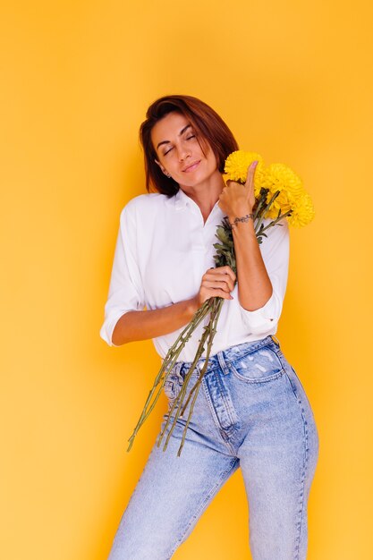 Studio tourné sur fond jaune heureux femme caucasienne cheveux courts portant des vêtements décontractés chemise blanche et pantalon en jean tenant bouquet d'asters jaunes