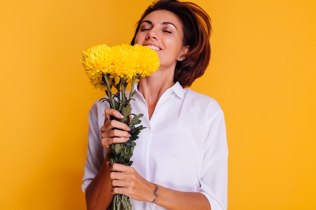 Studio tourné sur fond jaune heureux femme caucasienne cheveux courts portant des vêtements décontractés chemise blanche et pantalon en jean tenant bouquet d'asters jaunes