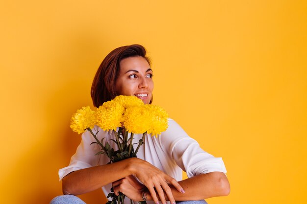 Studio tourné sur fond jaune heureux femme caucasienne cheveux courts portant des vêtements décontractés chemise blanche et pantalon en jean tenant bouquet d'asters jaunes