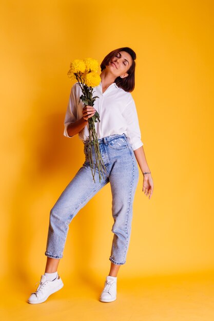 Studio tourné sur fond jaune heureux femme caucasienne cheveux courts portant des vêtements décontractés chemise blanche et pantalon en jean tenant bouquet d'asters jaunes