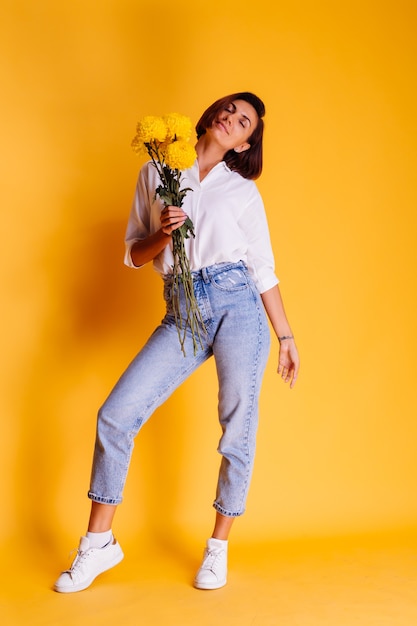 Studio tourné sur fond jaune heureux femme caucasienne cheveux courts portant des vêtements décontractés chemise blanche et pantalon en jean tenant bouquet d'asters jaunes