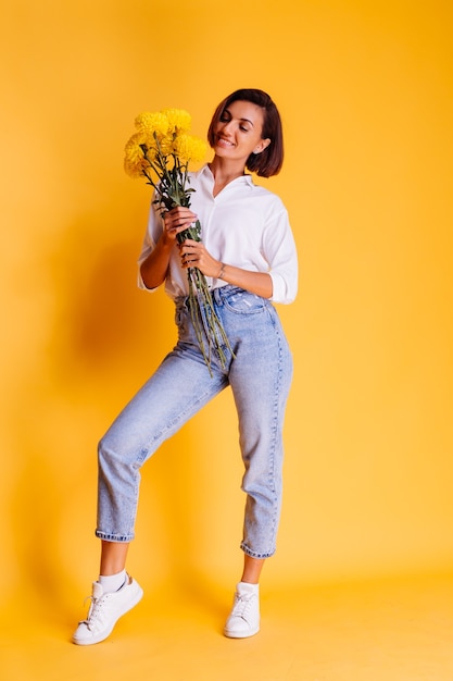 Studio tourné sur fond jaune heureux femme caucasienne cheveux courts portant des vêtements décontractés chemise blanche et pantalon en jean tenant bouquet d'asters jaunes