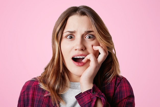 Photo gratuite stupéfaite, une jolie femme émotive regarde la caméra, garde la bouche ouverte, mord les ongles comme des soucis, étant choquée de réaliser des nouvelles horrifiées