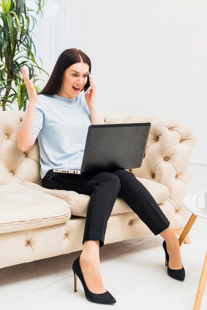 Stupéfié jeune femme assise sur un canapé avec ordinateur portable et parler par téléphone