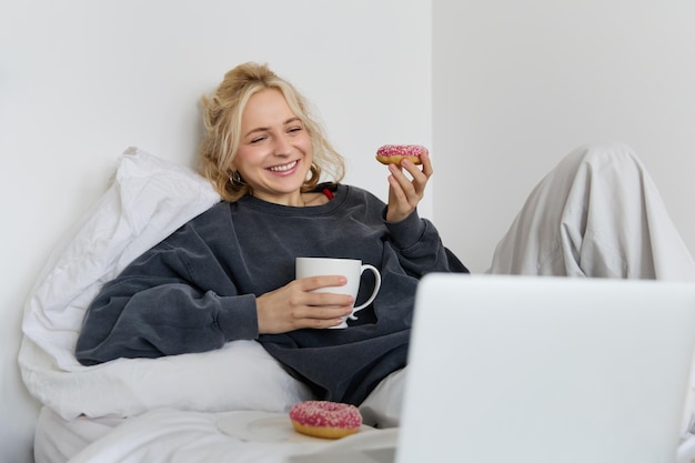 Photo gratuite style de vie et concept de personnes femme blonde heureuse allongée dans le lit avec de la nourriture buvant du thé et mangeant