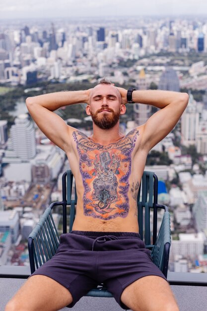 Succès bel homme fort tatoué brutal barbu européen aux seins nus avec montre est assis sur une chaise à l'étage élevé avec vue imprenable sur la ville