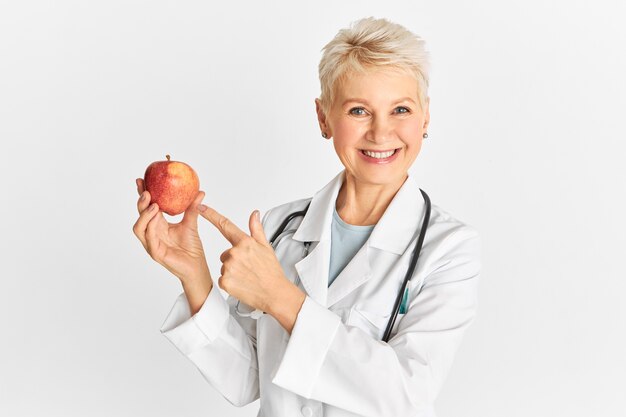 Succès femme médecin d'âge moyen portant un uniforme médical souriant à la caméra et pointant le doigt sur la pomme rouge mûre qui est bonne pour la santé intestinale et favorise la perte de poids. Santé et alimentation