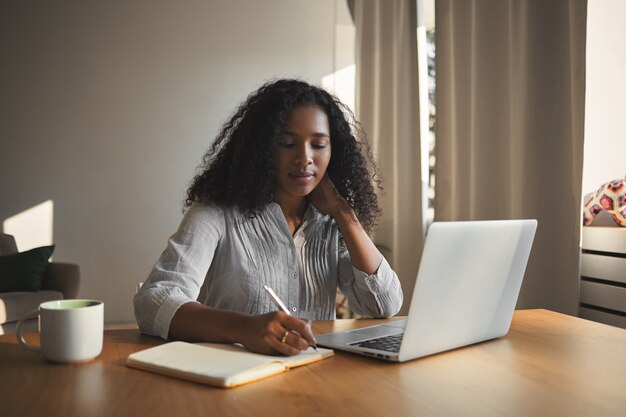 Succès séduisante jeune femme d'affaires afro-américaine en chemise élégante assis sur son lieu de travail devant un ordinateur portable ouvert et prendre des notes dans son journal, ayant une expression faciale réfléchie