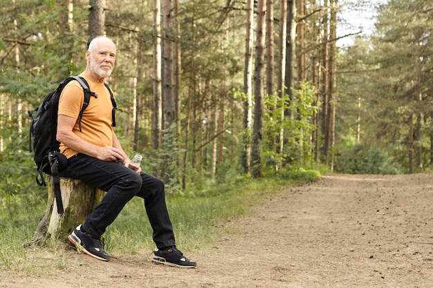 Summertime, Randonnée, Mode De Vie Actif Et Concept D'âge. Homme De Race Blanche énergique à La Retraite, Passer La Journée D'été à L'extérieur Dans La Nature Sauvage, Voyager à Pied, Se Reposer Sur Une Souche Avec Une Bouteille D'eau