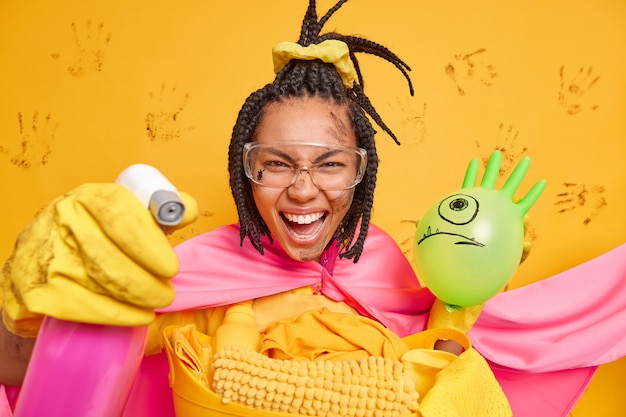 Photo gratuite une super-femme joyeuse dans des gants en caoutchouc de manteau rose, des lunettes de protection, tient une bouteille de distribution et un ballon gonflé nettoie toutes les pièces de la maison isolées sur un mur jaune