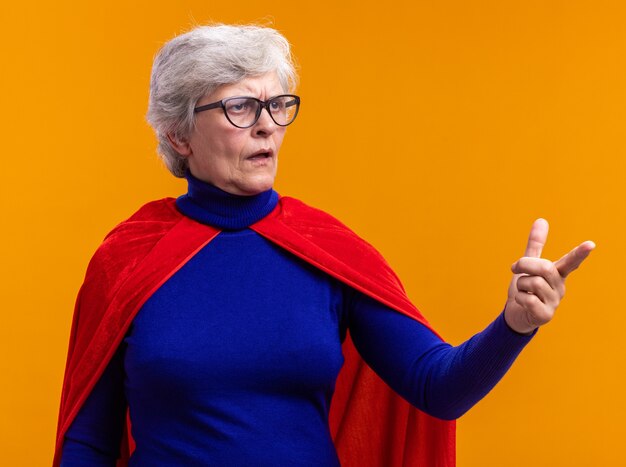 Super-héros femme senior avec des lunettes portant une cape rouge regardant de côté avec une expression confuse pointant