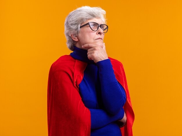 Super-héros femme senior avec des lunettes portant une cape rouge regardant de côté avec une expression pensive pensant debout sur fond orange