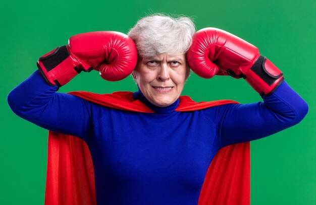 Super-héros femme senior portant une cape rouge avec des gants de boxe levant les yeux en étant agacé et irrité debout sur fond vert