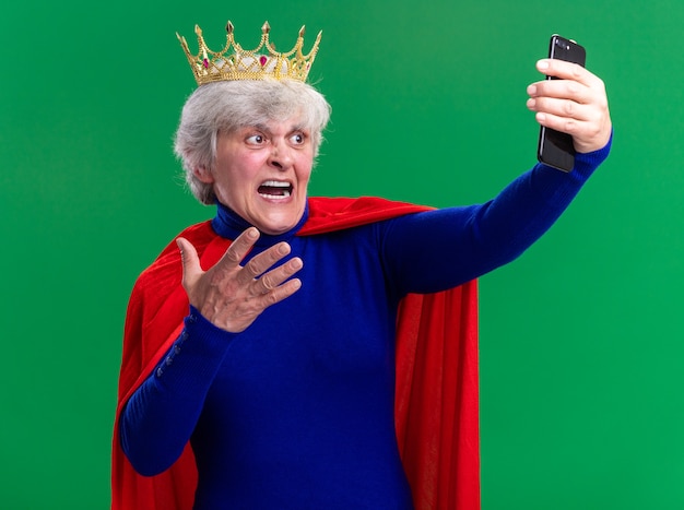 Photo gratuite super-héros femme senior portant une cape rouge et des lunettes avec une couronne sur la tête à l'aide d'un smartphone regardant l'écran avec une expression agressive criant debout sur fond vert