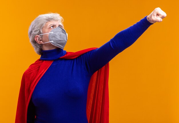 Super-héros femme senior portant une cape rouge et un masque de protection faciale faisant le geste du gagnant