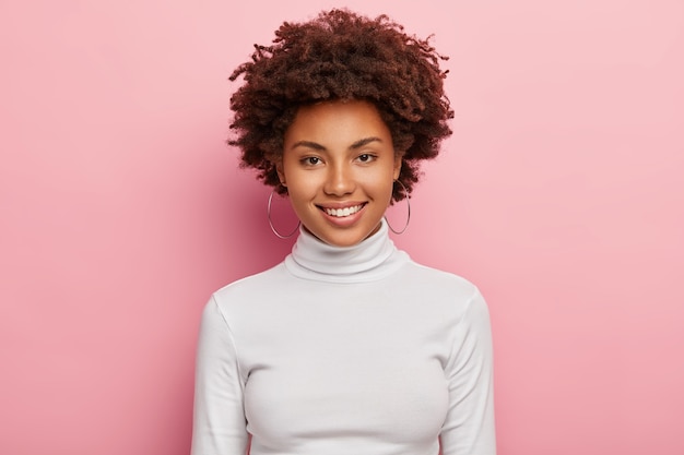 Photo gratuite superbe belle femme a un sourire agréable sur le visage, une peau saine, une coupe de cheveux afro, porte un pull à col roulé blanc décontracté