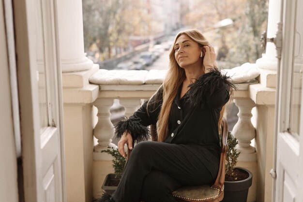 Superbe blonde caucasienne adulte en pyjama noir en soie utilise des écouteurs assis sur le balcon pendant la journée Mode de vie de loisirs et concept de beauté