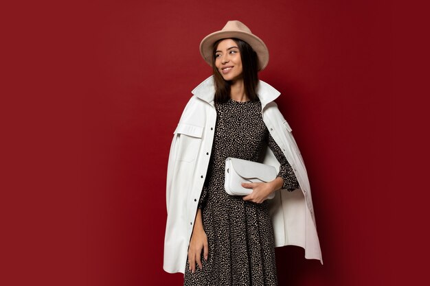 superbe brune européenne gir en veste blanche à la mode et robe avec impression posant. Tenant le sac à main en cuir.