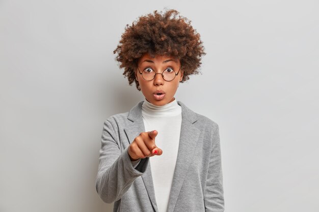 Une superbe femme d'affaires aux cheveux bouclés impressionnée pointe directement avec l'index, voit quelque chose d'étonnant devant, porte une tenue formelle