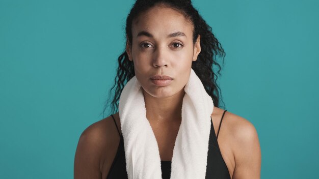 Superbe femme afro avec une serviette sur l'épaule regardant directement à huis clos fatiguée après un entraînement actif sur fond bleu Concept de sport et d'exercice